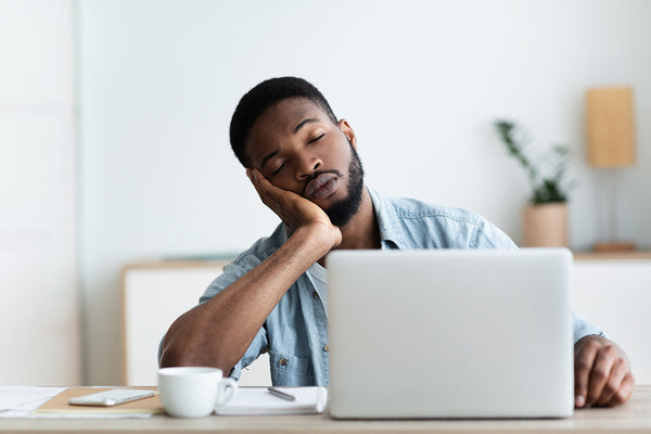 Man asleep at desk with laptop and coffee cup, feeling tired.