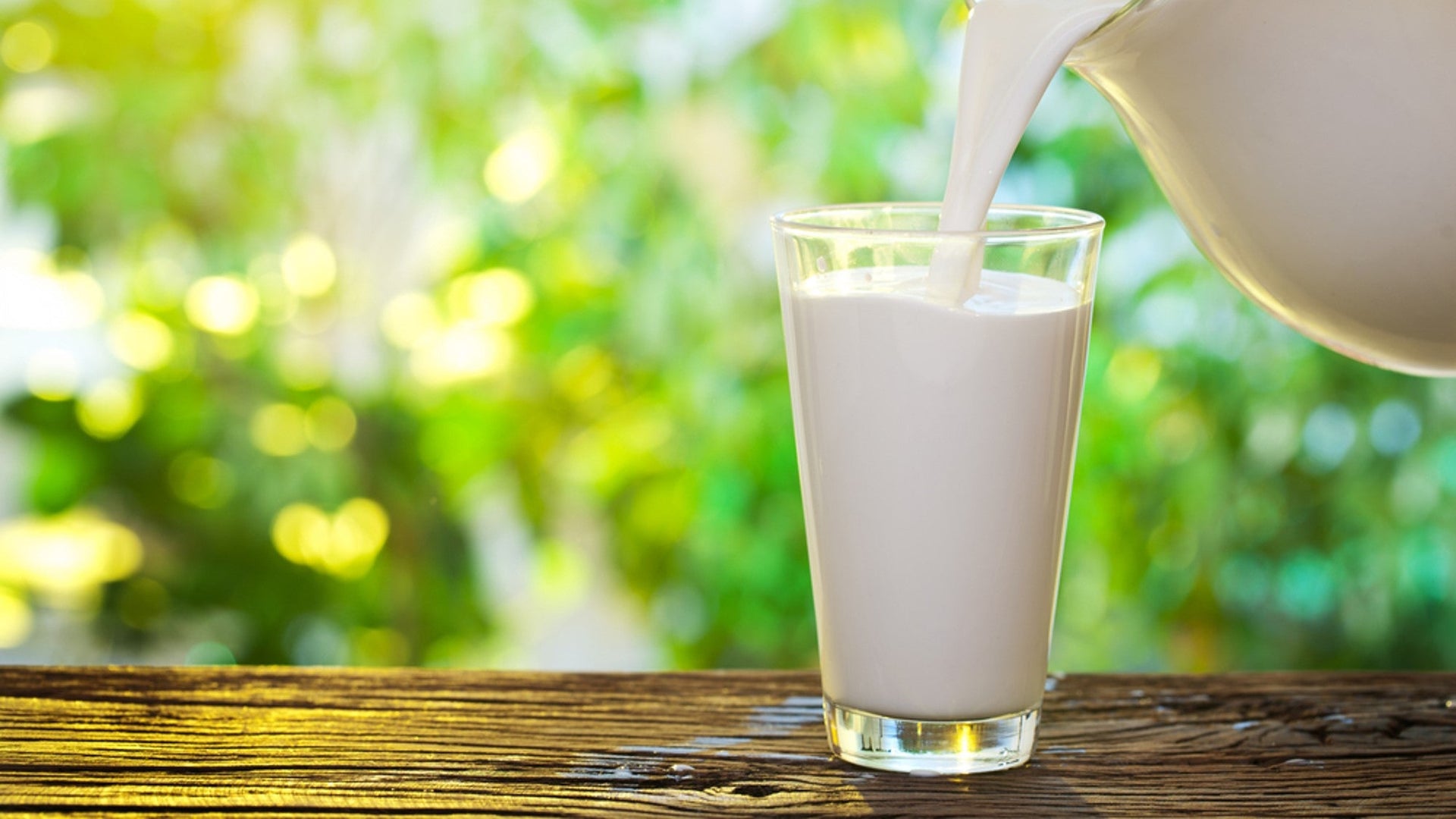 Pouring fresh milk into glass on wooden table outdoors.