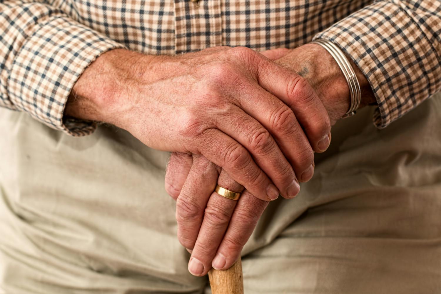 Elderly man's hands resting on a cane, wearing a checkered shirt.