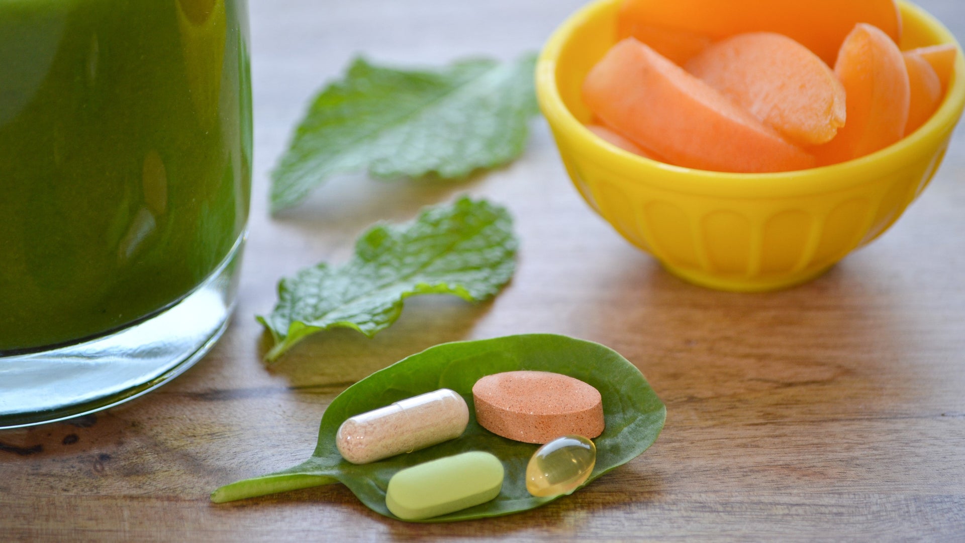 Green smoothie with supplements on spinach leaf, bowl of cantaloupe slices nearby.